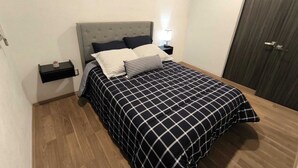 A neatly made bed with a checkered bedspread, a bedside table with a lamp, and a small shelf with a plant. - Modern and centrally located apartment in CDMX (Ciudad de México)