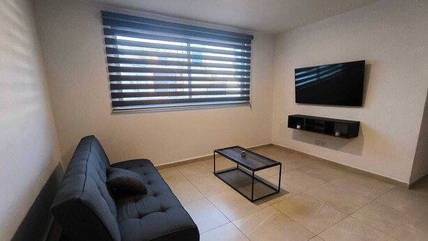 A modern living room with a dark sofa, a coffee table, a TV mounted on the wall, and a window with blinds. - Modern and centrally located apartment in CDMX (Ciudad de México)