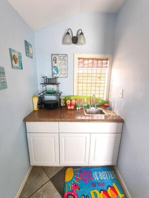 A small kitchen with white cabinets, a window with blinds, and a colorful rug on the floor.