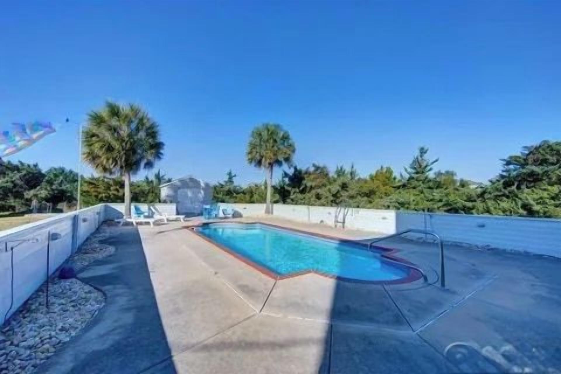 Una piscina con agua cristalina azul, rodeada de una terraza de concreto, palmeras y un cielo despejado.