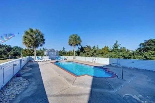 A swimming pool with a clear blue water, surrounded by a concrete deck, palm trees, and a clear sky.