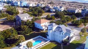 Vista aérea de una zona residencial costera con casas, una casa grande de color blanco con piscina, y una playa al fondo.