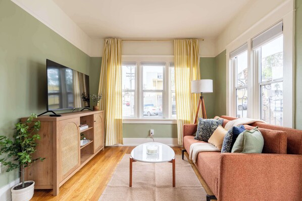 Ein Wohnzimmer mit Sofa, Couchtisch, Fernseher auf einem Holzschrank und einem Fenster mit Vorhängen.