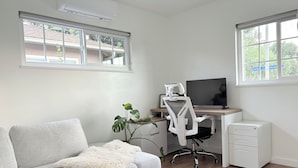 Un espacio de oficina moderno con un sofá de gris claro, un escritorio blanco con computadora y monitor, una silla de oficina blanca y un archivador pequeño de color blanco.