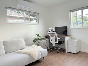 Ein moderner Arbeitsbereich mit einem hellgrauen Sofa, einem weißen Schreibtisch mit Computer und Bildschirm, einem weißen Bürostuhl und einer kleinen weißen Aktenschrank.