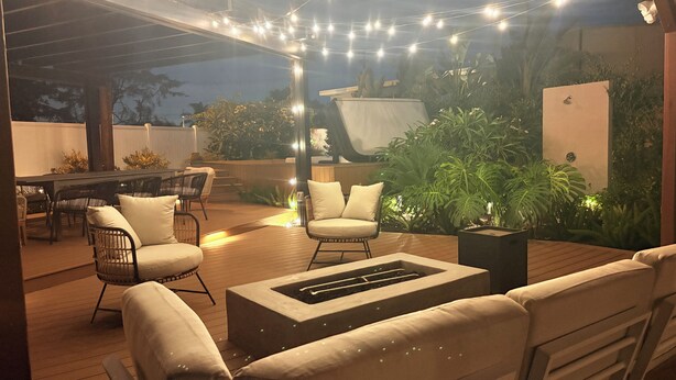 Un patio exterior con suelo de madera, asientos acolchados, un pozo de fuego, luces colgantes y abundante vegetación.