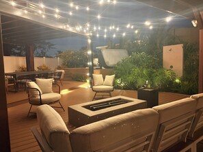 An outdoor patio with wooden flooring, cushioned seating, a fire pit, string lights, and lush greenery. - Mid-century modern home with huge backyard + hot tub (Long Beach)