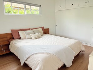 Ein ordentlich bezogenes Bett mit weiß-beigen Farben, ein hölzernes Kopfende und ein Fenster mit Blick auf Grünflächen.