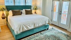 A bedroom with a teal bed, white bedding, leopard print pillows, two bedside lamps, a floor lamp, a patterned rug, and a balcony with a white railing. - Coastal Retreat Minutes to Sullivans Island and downtown Charleston (Mount Pleasant)