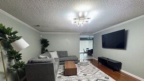 Een woonkamer met een grijze hoekbank, een houten salontafel, een kamerkleed met een patroon, een vloerlamp en een flatscreen televisie.