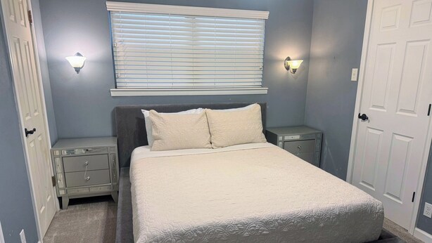Een slaapkamer met een bed, een raam met zonwering, twee nachtkastjes en een deur.