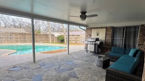 Een overdekt terras met stenen vloer, een zwembad, een barbecue en een zithoek.