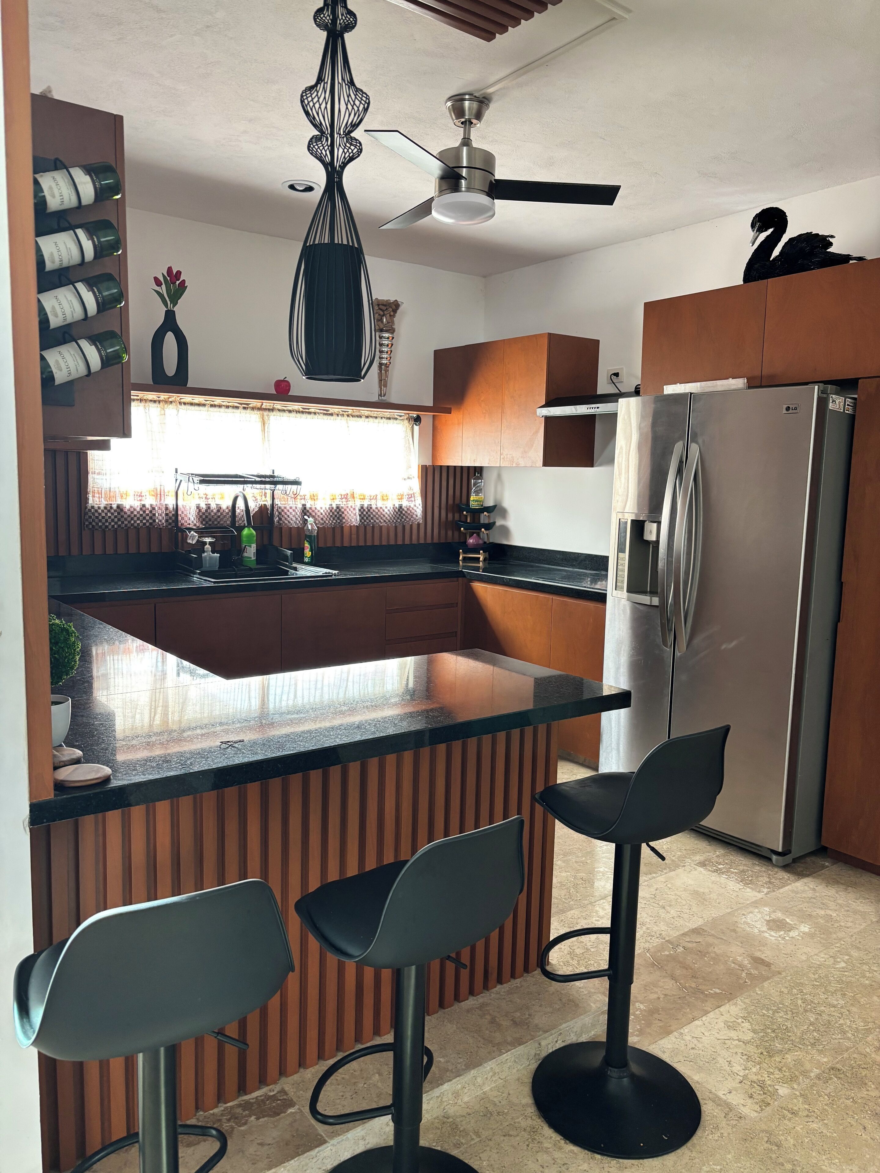 Une cuisine moderne avec des meubles en bois, un réfrigérateur en acier inoxydable, un coin bar avec des tabourets et un ventilateur de plafond.