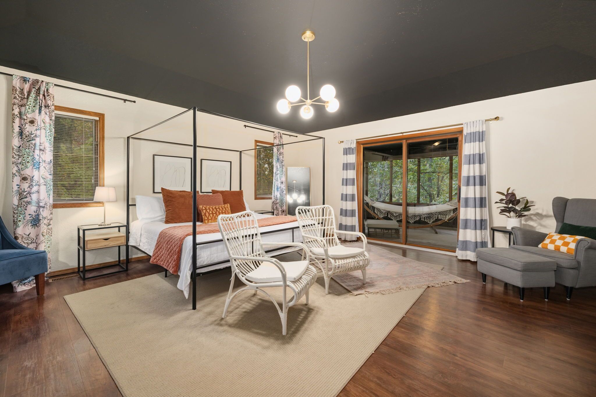 A bedroom with a four-poster bed, two white wicker chairs, a grey sofa, and a large window with a view of greenery.