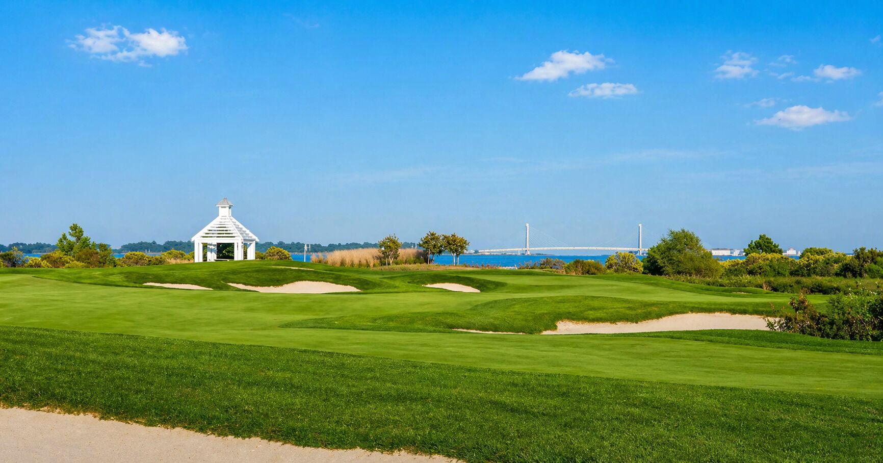 A golf course with sand bunkers, a white pavilion, and a bridge in the background.