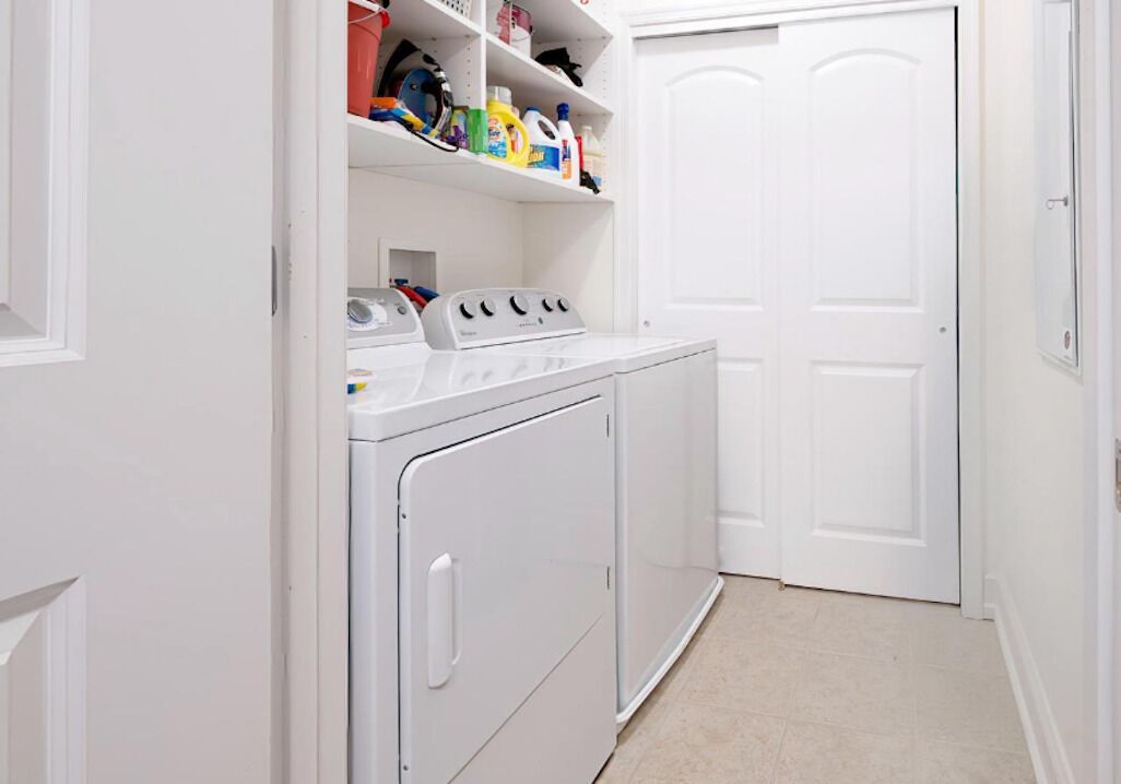 A laundry room with a front-loading washer and dryer, shelves with cleaning supplies, and a white door in the background.