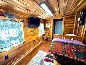 A wooden cabin room with a bed, a television, a chair, and a window with coffee-themed curtains. - Charming Lakeside Cabin on Lookout mountain, Fort Payne by Little River Canyon! (Fort Payne)