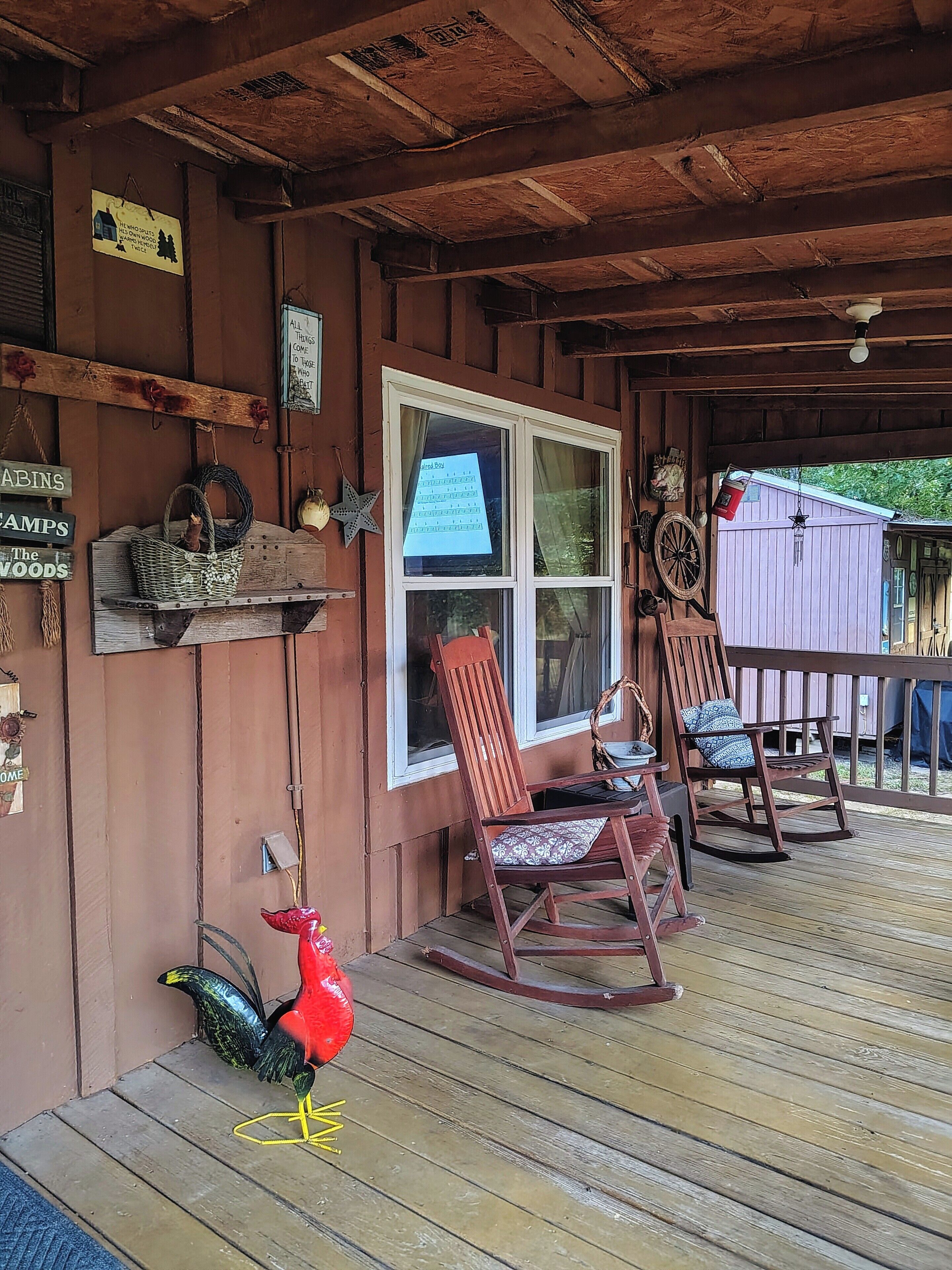 A wooden porch with two rocking chairs, a wooden table, and a window with a view of a shed.