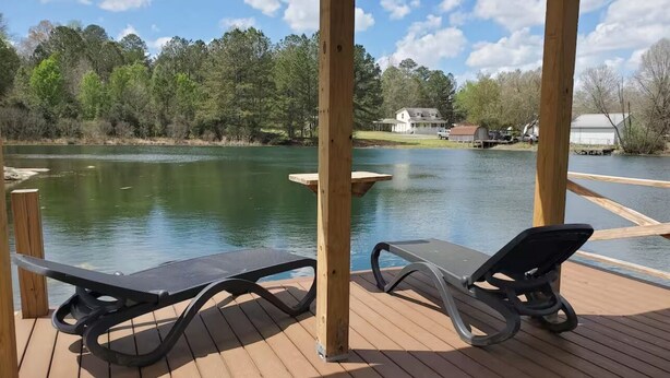 Une terrasse en bois avec deux chaises longues donnant sur un lac, entourée d’arbres et avec une maison en arrière-plan.