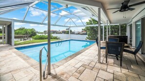 A covered outdoor swimming pool with a glass ceiling, a ceiling fan, and a view of a body of water and buildings in the background. - Charming 3-bedroom waterfront house in Naples/Marco Island (Naples)