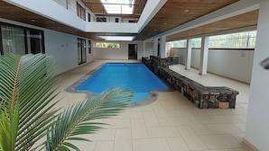 A modern indoor swimming pool with a stone deck, surrounded by a multi-level building with wooden beams and large windows. - Modern 2-Bedroom Apartment Near La Croisette, Grand Baie (Grand Baie Heights)