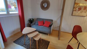 Un espacio habitable pequeño con un sofá gris, cojines rojos, una mesita y un espejo colgado en la pared.