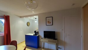 Una sala moderna con un sofá azul, una mesa de comedor, un televisor en un mueble bajo, una cafetera y un ventanal con cortinas rojas.