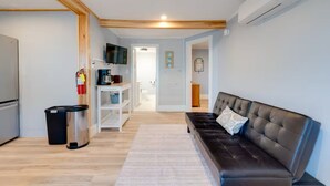 A modern living room with a sofa, a coffee table, and a TV. There is a kitchen area with a fridge, a coffee machine, and a trash bin. The room has wooden beams on the ceiling and a white rug on the floor. - Charming Beachfront Loft | New Renovation (Old Orchard Beach)