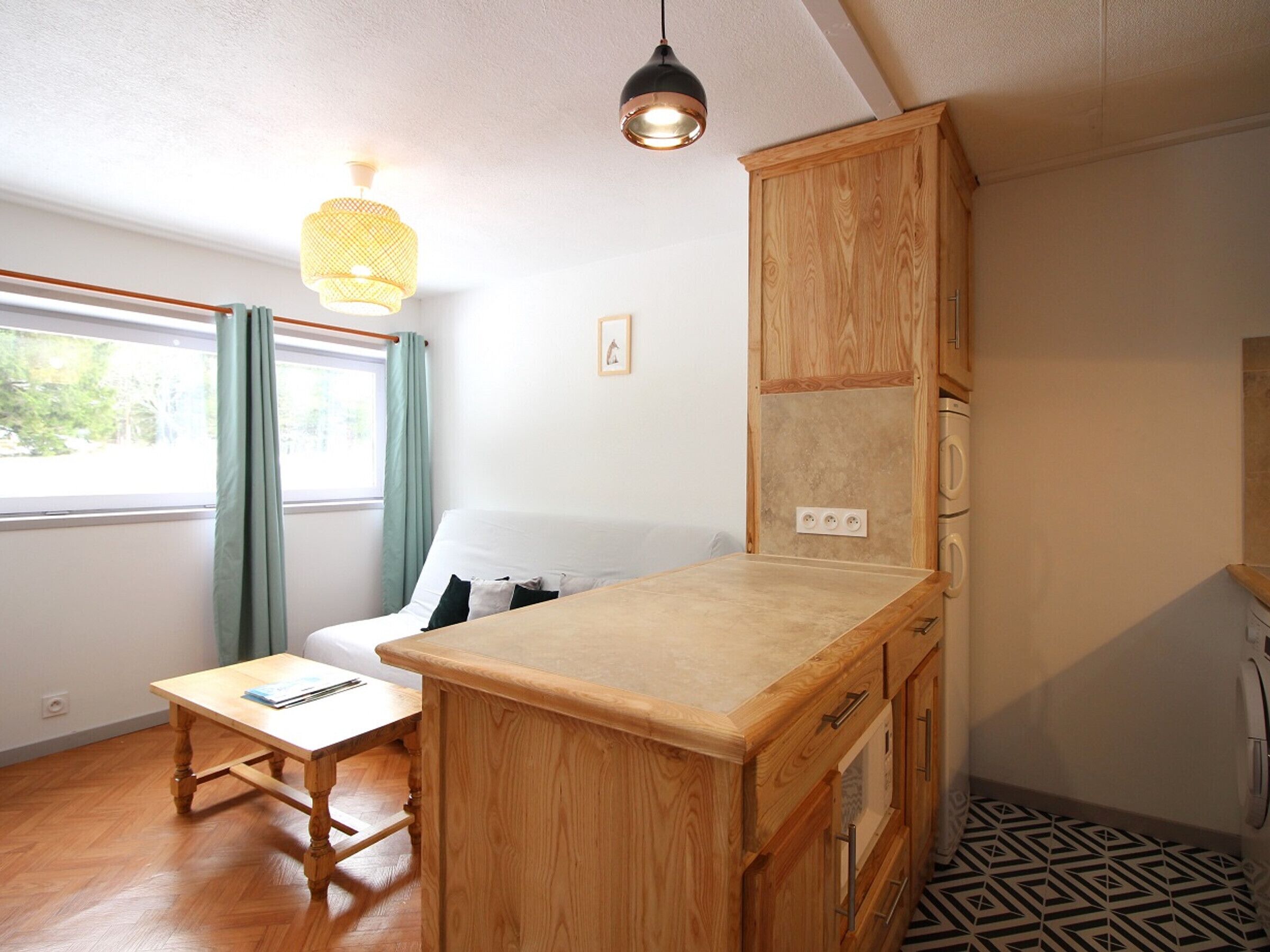 A compact room with a wooden desk, a small table, a kitchenette with a built-in oven, and a window with curtains.