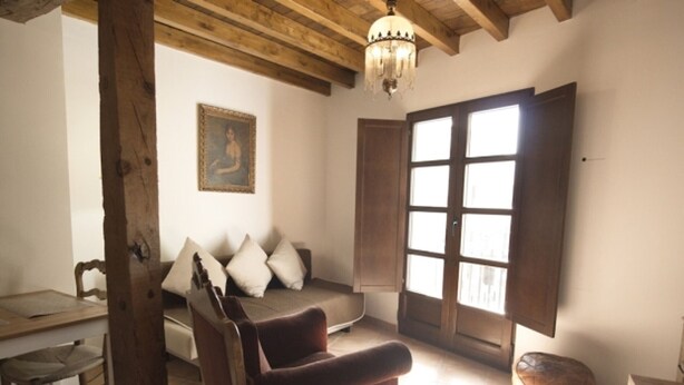 A living room with a wooden ceiling, a sofa with cushions, a wooden chair, a table with a chair, and a large window with shutters. - Historic Studio in Granada, Near Alhambra (Granada)