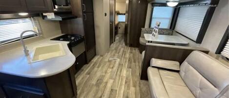 A compact RV interior with a kitchenette, a sink, a stove, a microwave, and a seating area with a window.