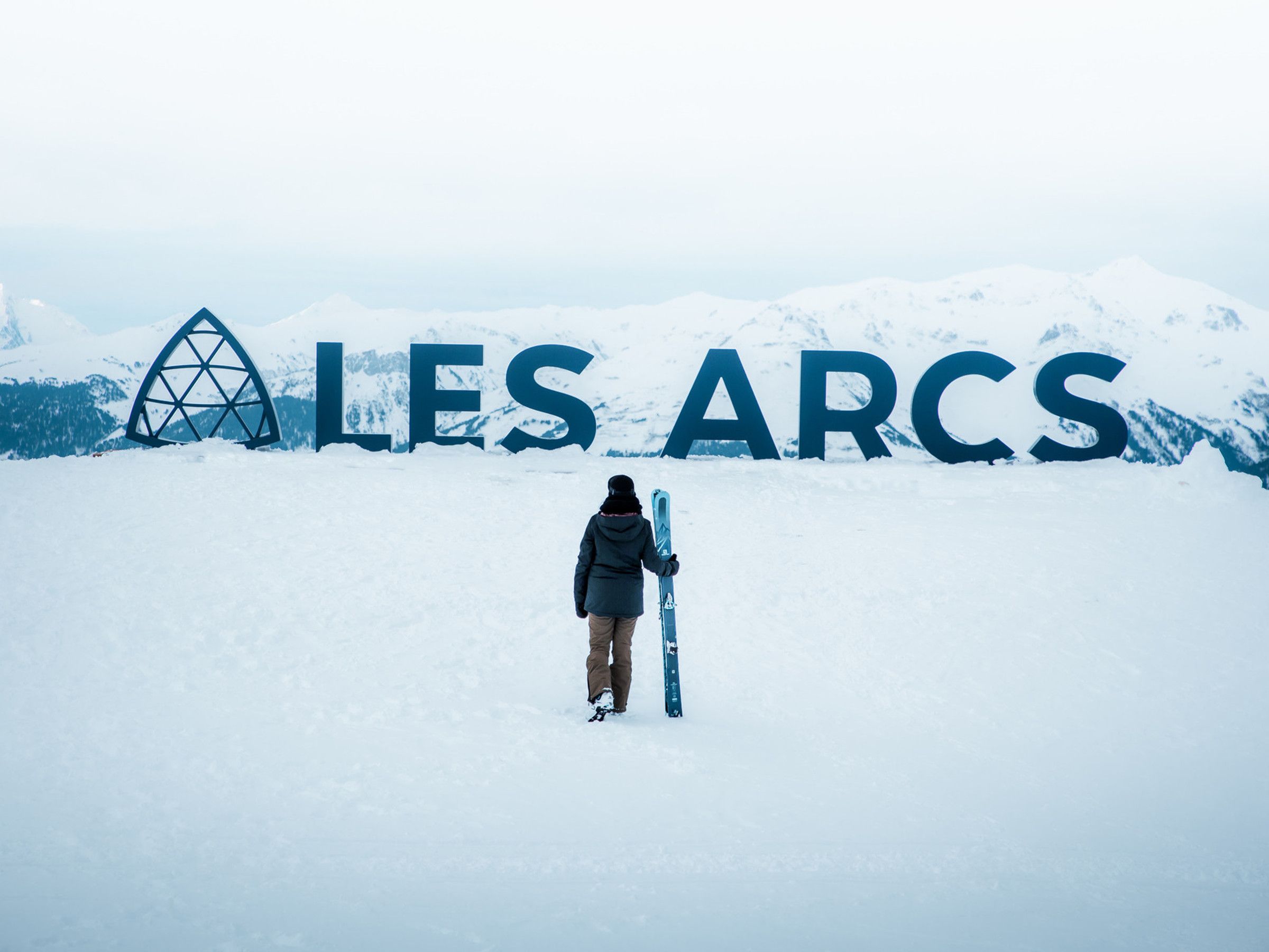 Eine Person steht vor einem großen, mit Schnee bedeckten Berg, auf dem der Schriftzug 'LES ARCS' zu sehen ist.