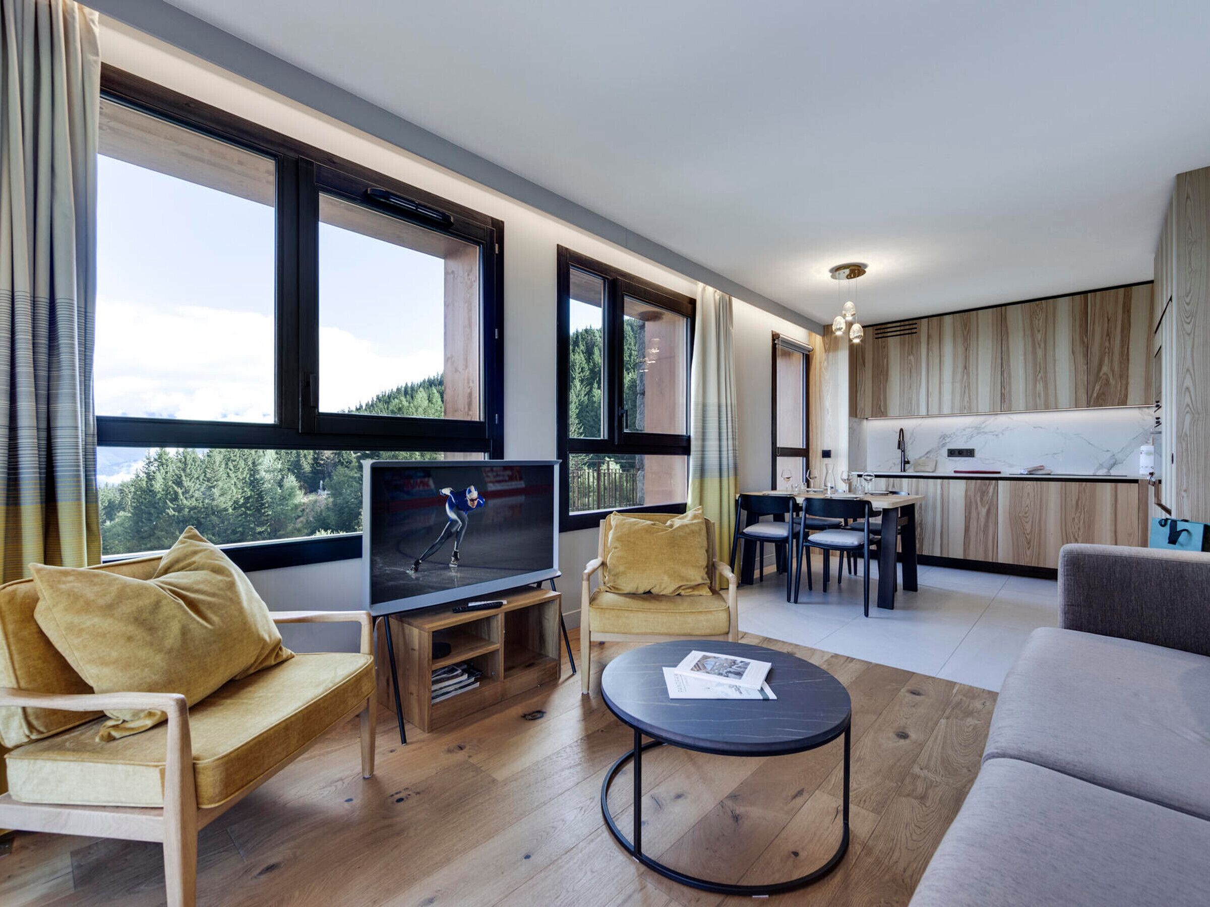 Een moderne woonkamer met een televisie, bank en salontafel. Er is een eethoek met stoelen en een keuken op de achtergrond.