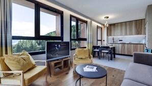 Een moderne woonkamer met een televisie, bank en salontafel. Er is een eethoek met stoelen en een keuken op de achtergrond.
