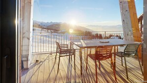 Eine Holzterrasse mit Tisch und Stühlen, mit Blick auf eine verschneite Landschaft mit Bergen und blauem Himmel.