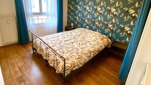 Une pièce avec un lit, un radiateur, une fenêtre avec des rideaux et un mur orné d’un motif de feuilles.