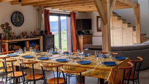 Zona pranzo con un lungo tavolo apparecchiato per un pasto, travi in legno al soffitto, un orologio a parete e vista sull'esterno attraverso una porta in vetro.