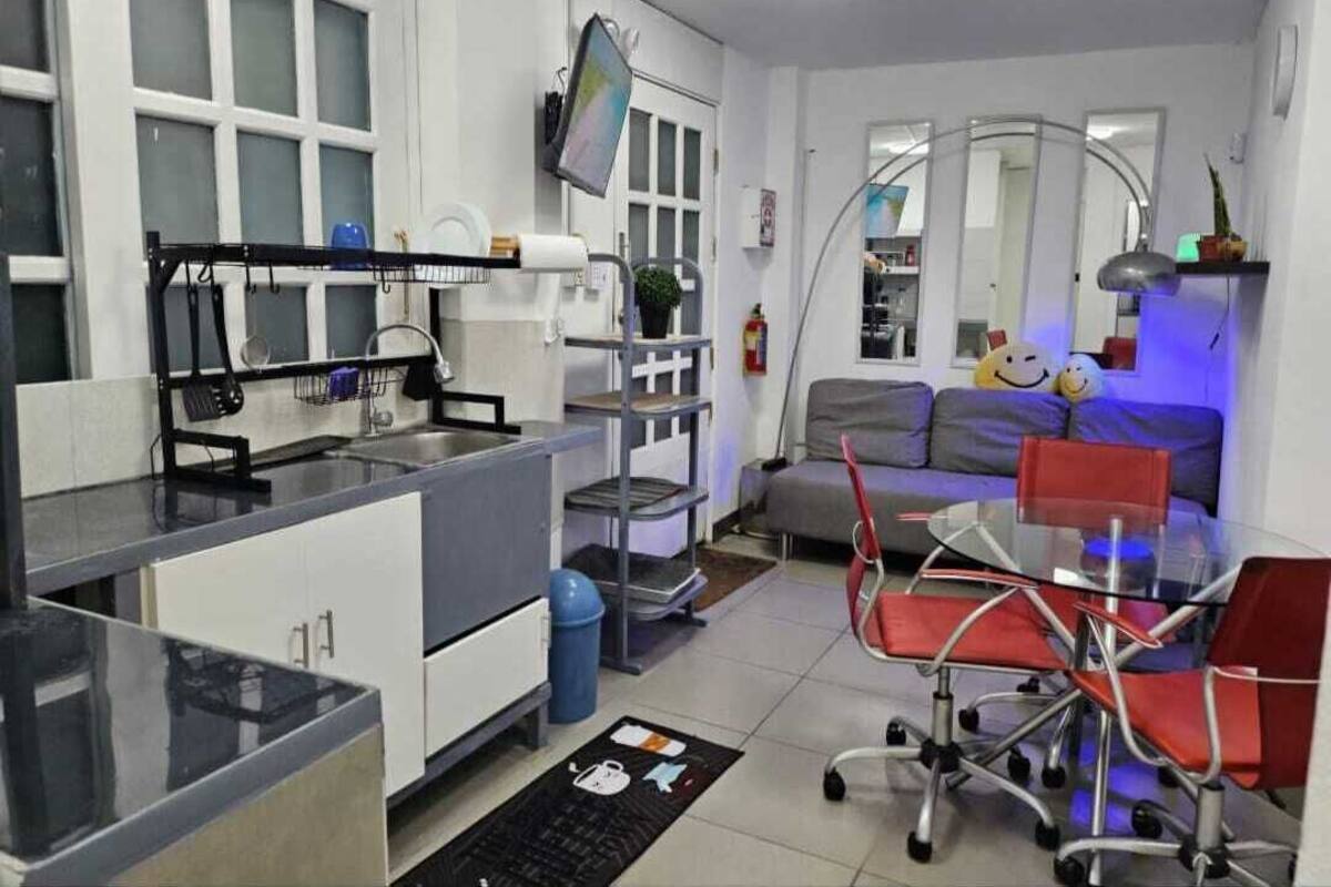 A compact living space with a kitchenette, a dining area with a glass table and red chairs, a sofa with cushions, and a large mirror.