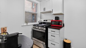A compact kitchen with white cabinets, a black stove, a red microwave, and a window with a view of a brick building.