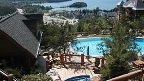 A resort with a large pool, wooden deck, and a view of a lake and buildings in the background. - Two Apartment 152/2+152/3 Ski In/Ski out (Mont-Tremblant)