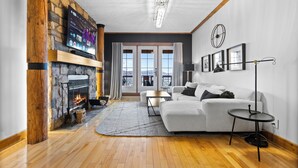 A modern living room with a stone fireplace, a white sofa, and a large TV mounted on a wooden wall. - Equinoxe 152-2 Ski in/ski out (Mont-Tremblant)