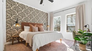 Quarto com papel de parede estampado, cama com várias almofadas, ventoinha no teto, janela com cortinas e planta em vaso.