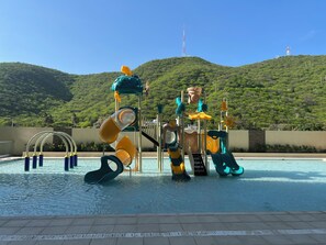 Un espace de jeux aquatiques avec des toboggans et des structures d’escalade, entouré d’une piscine offrant une vue sur des collines verdoyantes et un ciel dégagé.