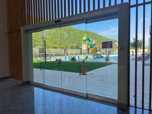 Vista attraverso una porta di vetro su un'area piscina con scivolo, struttura per arrampicata e un cartello che indica lavori in corso.