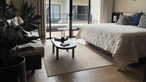 Et hotelværelse med en stor seng, en sofa, et lille bord og en potteplante. Der er en skydedør, der fører ud til en balkon med udsigt.