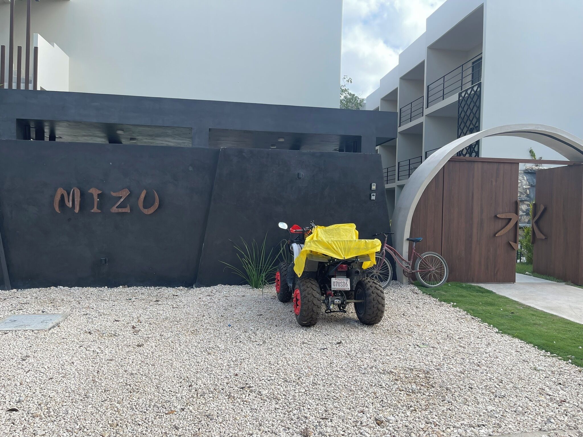 A gravel driveway leading to a modern building with a large black wall displaying the name "MIZU".