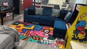 A room with a blue sofa, a colorful rug, a foosball table, a kitchenette, and a ceiling fan.