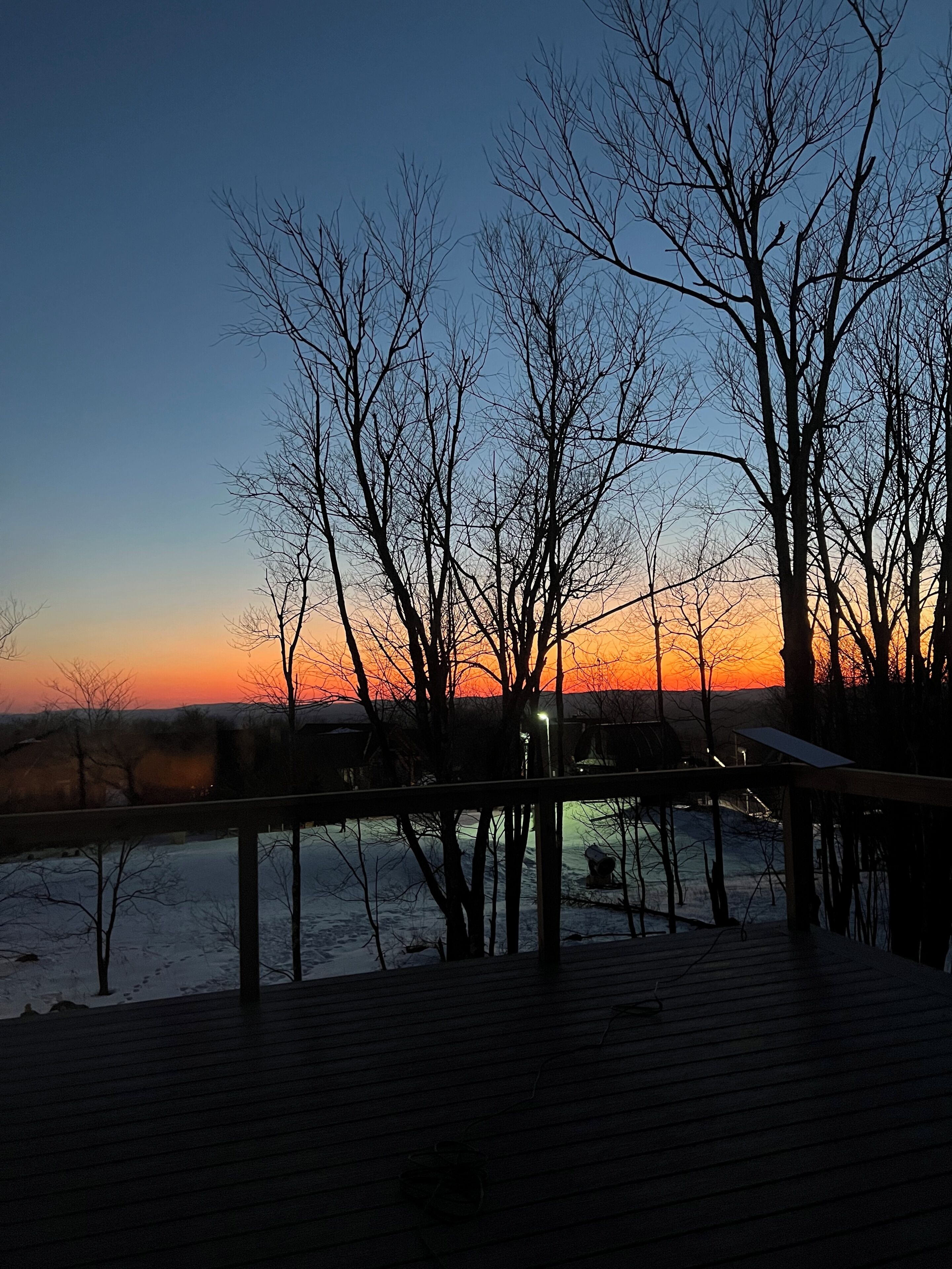 A wooden deck with a railing, overlooking a body of water and trees during sunset.