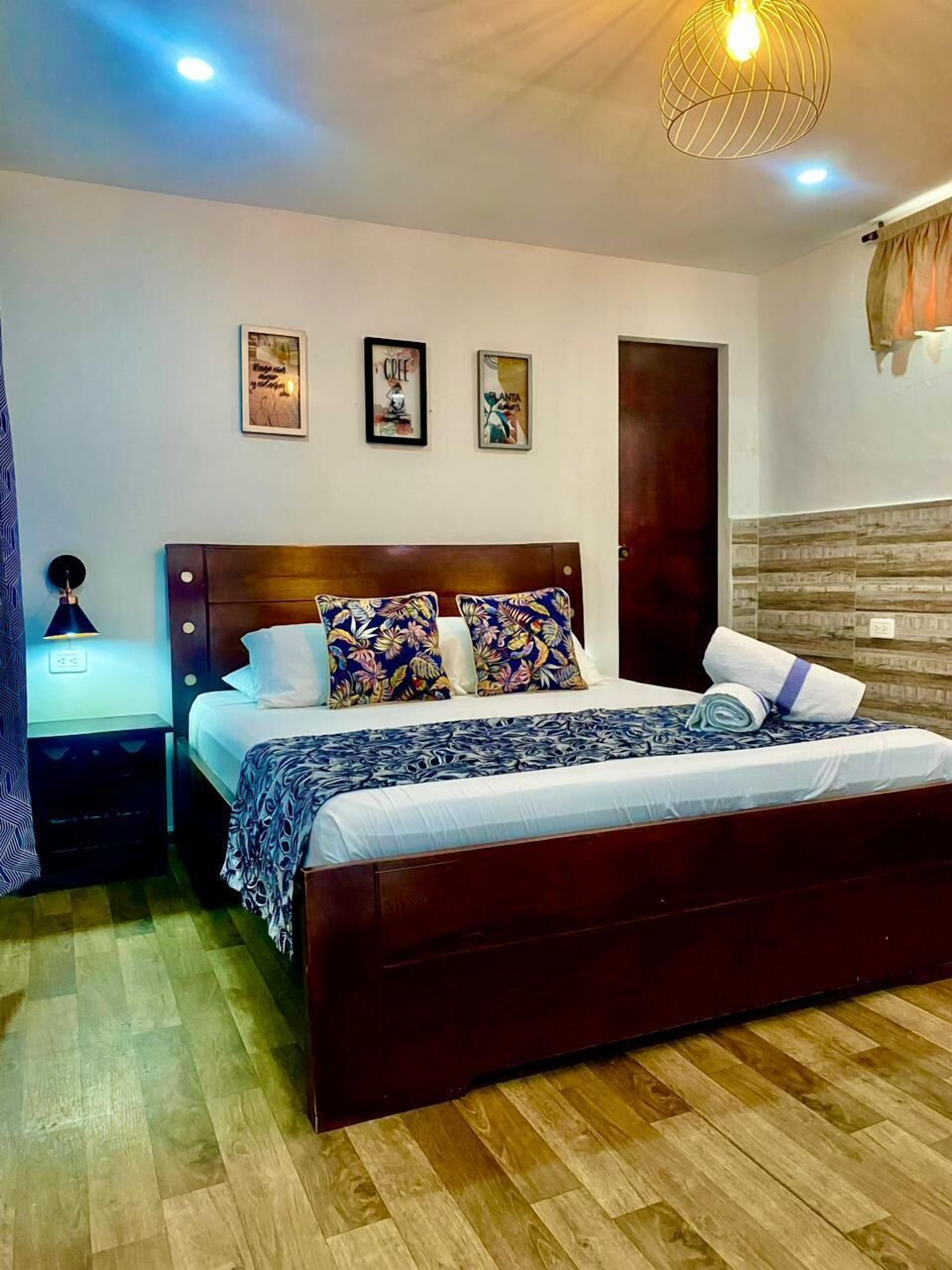 Un dormitorio con una cama de madera, una mesita de noche, una lámpara de pared y tres cuadros enmarcados.