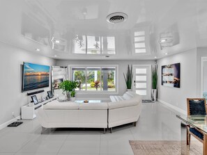 A spacious living room with a white sofa, a flat-screen TV, and a large window offering a view of the outdoors. - Lighthouse Point Retreat:Heated Pool & 3 Ensuite Bedrooms with luxury mattresses (Lighthouse Point)